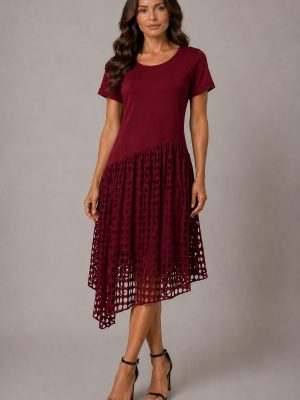 Elegant bordo rdeča obleka z asimetričnim spodnjim delom in dekorativnim izrezanim vzorcem. Model stoji v studijskem okolju v črnih sandalih z visoko peto, obleka pa poudarja ženstveno silhueto in lahkoten, sodoben videz.